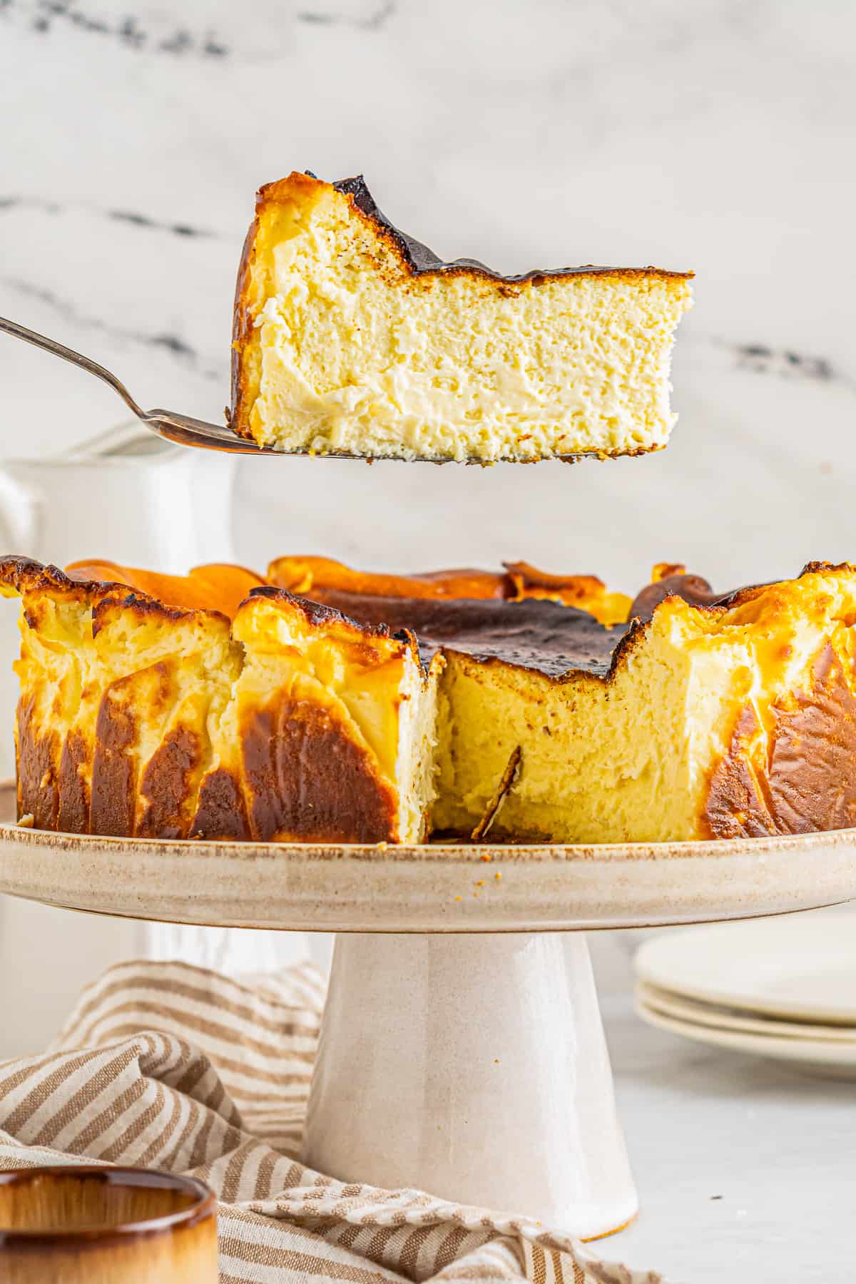 a slice of basque cheesecake on a cake server hovering above a cheesecake on a white cake stand.