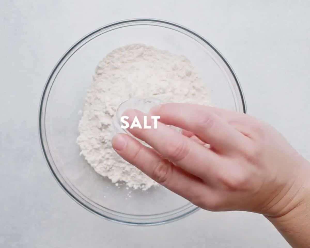 A hand adding salt to a glass bowl of whisked dry ingredients