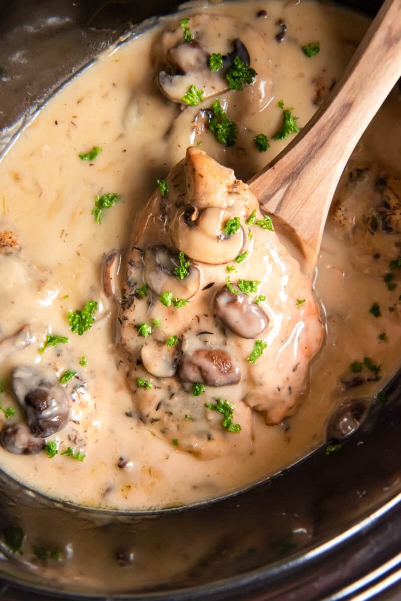 a wooden spoon lifting a chicken breast out of crockpot chicken marsala.