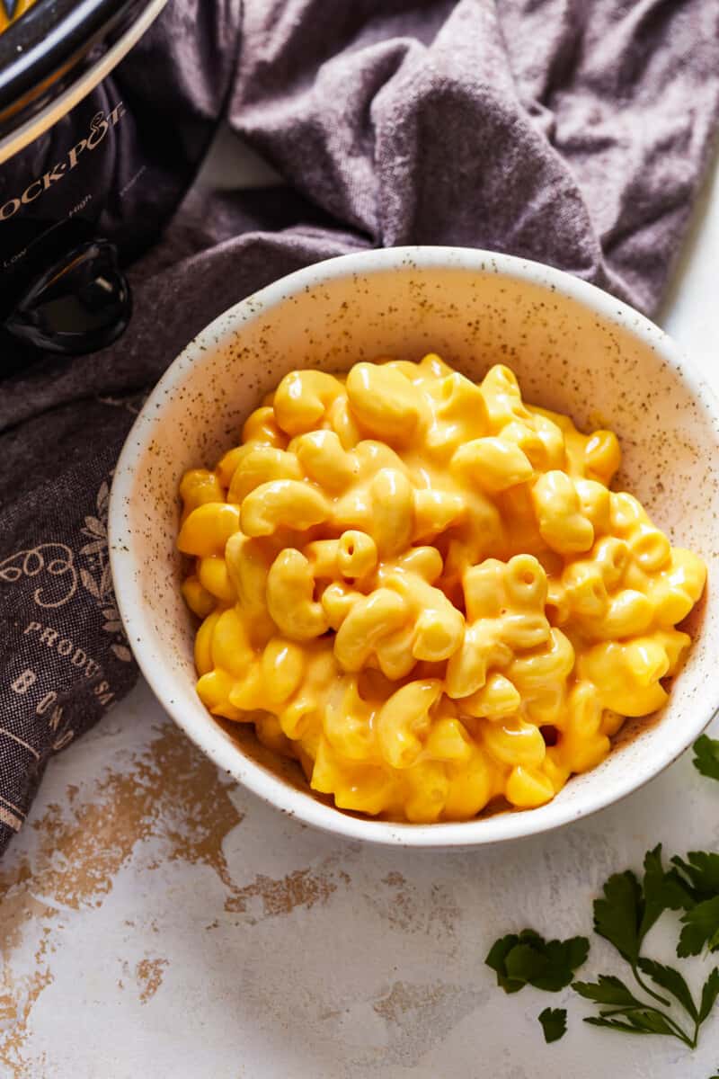 Crock Pot Mac and Cheese is a super simple recipe! This easy slow cooker macaroni and cheese makes the perfect side dish or weeknight dinner. It's virtually fool proof and comes out extra creamy and extra cheesy every time. Mac n cheese is always one of the best cheesy comfort foods around! a bowl of mac and cheese next to a crockpot