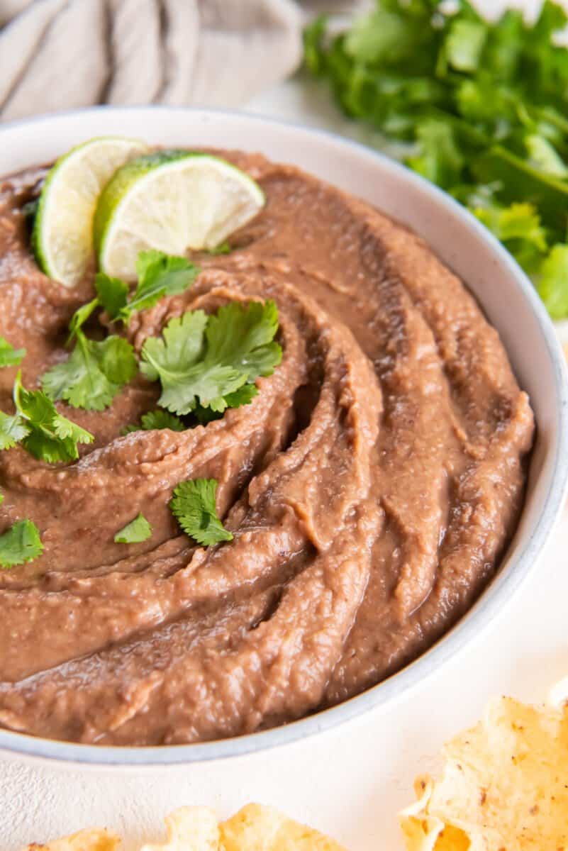 crockpot refried beans in a white bowl with cilantro and lime wedges.