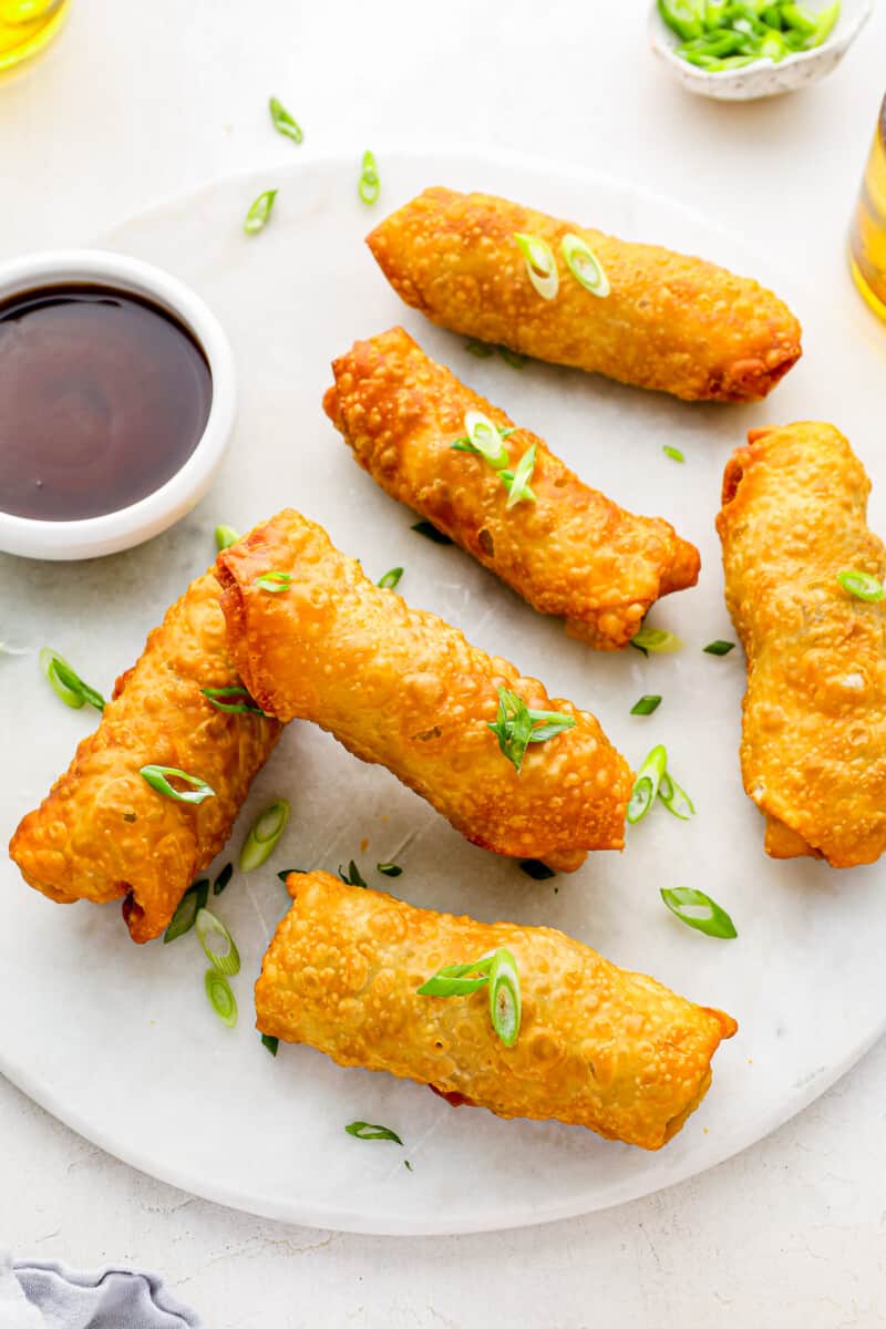 6 egg rolls on a white plate with dipping sauce.