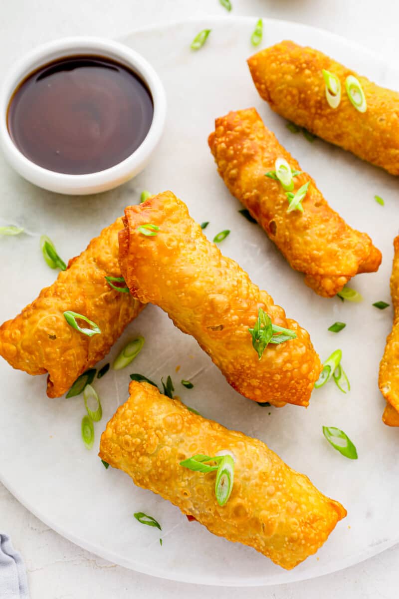 6 egg rolls on a white plate with dipping sauce.