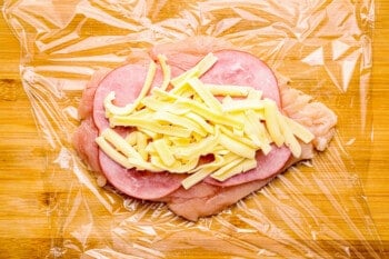 chicken layered with ham and cheese on plastic wrap.