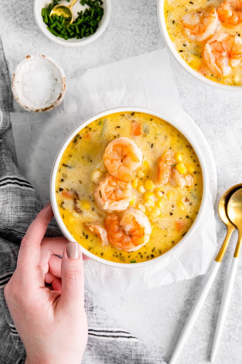 hand holding shrimp corn chowder in a white bowl with spoons.