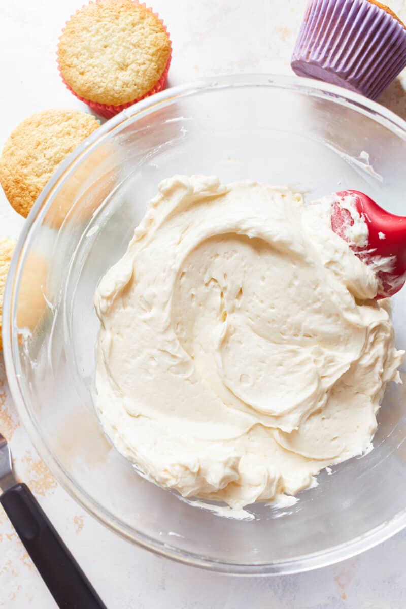 glass bowl filled with vanilla frosting, surrounded by unfrosted cupcakes