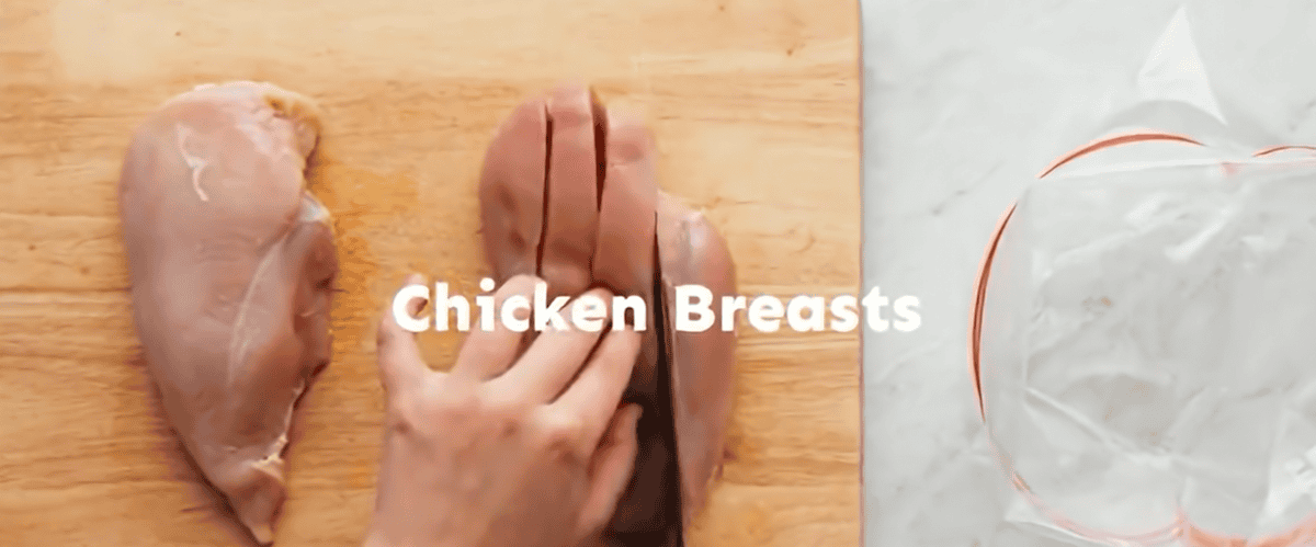 Slicing chicken breasts on a wooden cutting board with a plastic bag on the side.