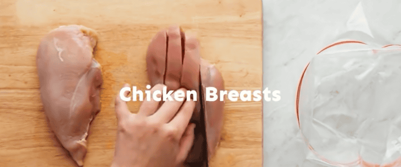 Slicing chicken breasts on a wooden cutting board with a plastic bag on the side.