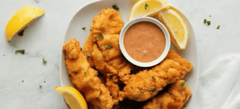 Chicken fingers on a plate with sauce and lemon wedges.