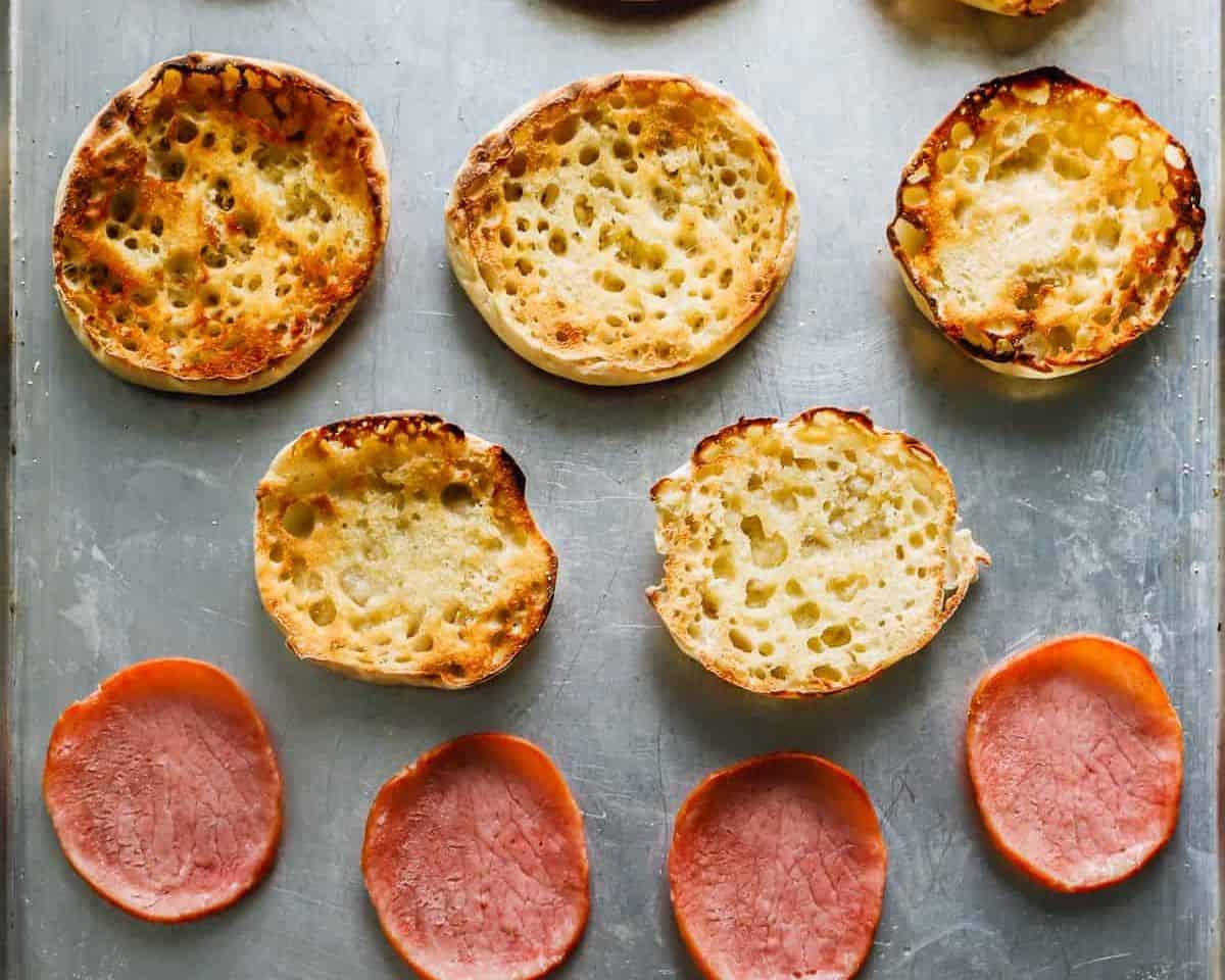 Toasted English muffins and Canadian bacon on a cookie sheet. 