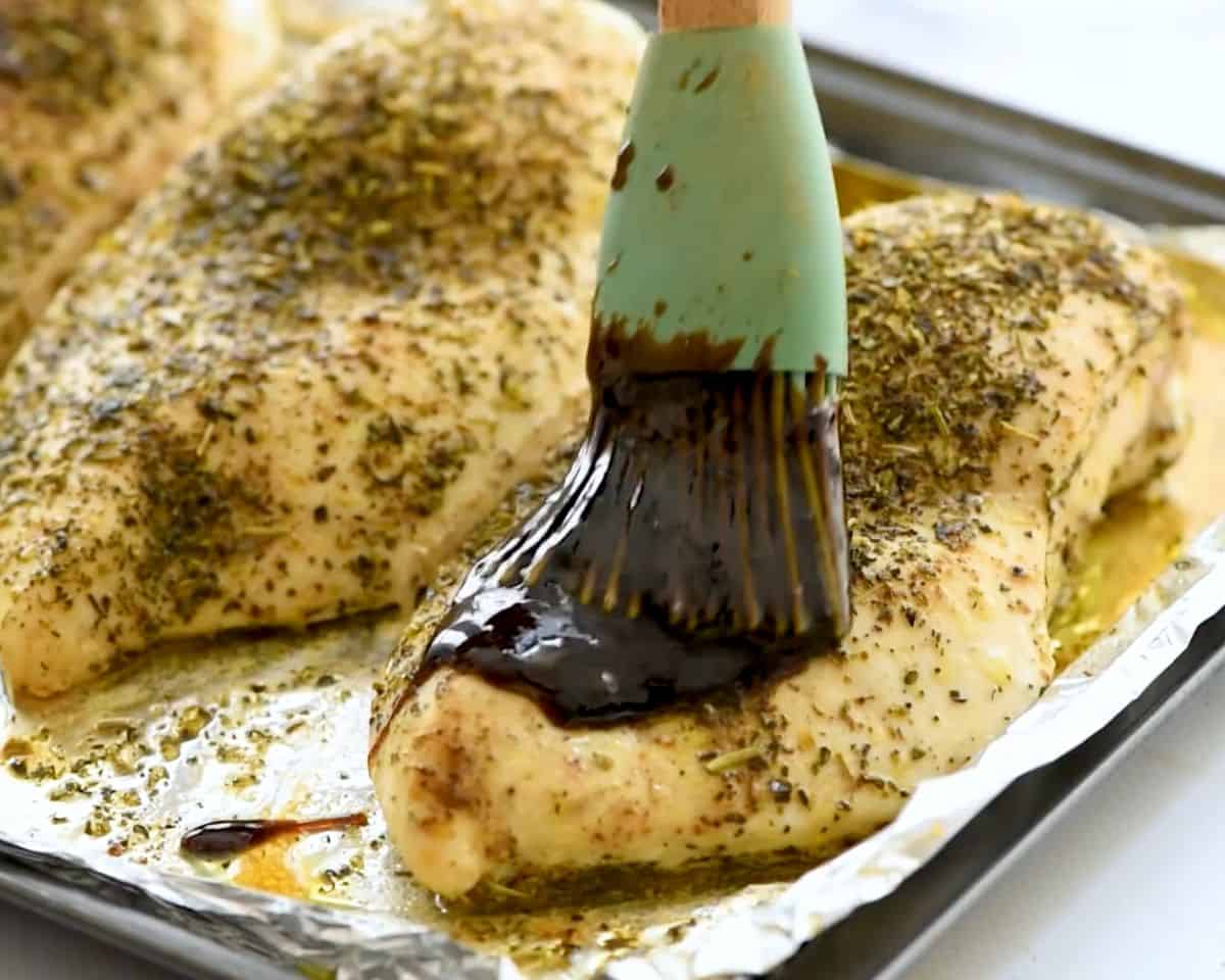 Brushing honey balsamic glaze onto seasoned chicken breasts on a baking sheet before finishing in the oven