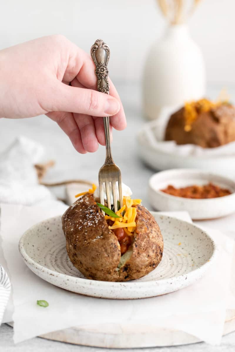 a hand holding a fork piercing a loaded air fryer baked potato on a white plate.