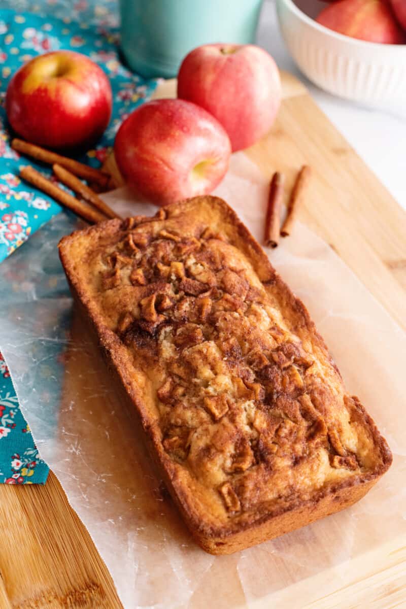 fresh baked loaf of apple cinnamon bread on a cutting board, with apples and cinnamon sticks scattered around