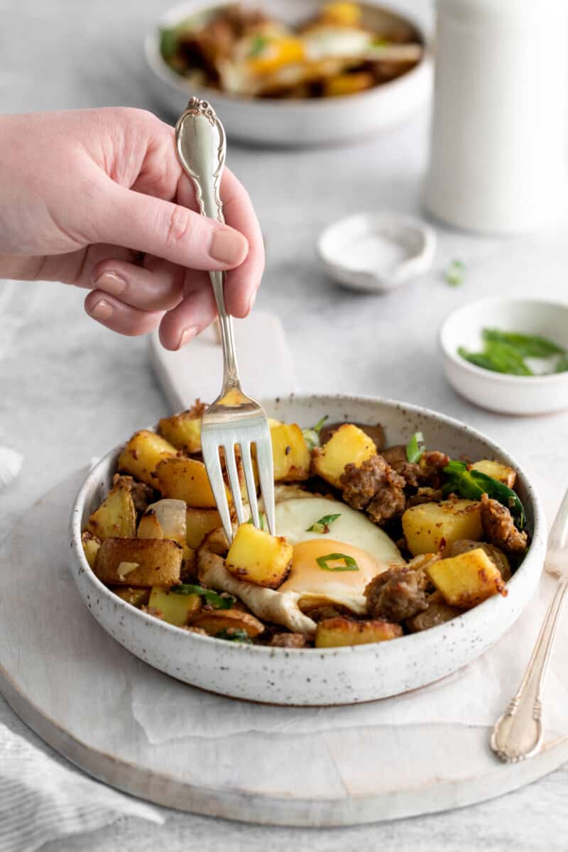 a fork stabbing a potato from breakfast hash in a white bowl.