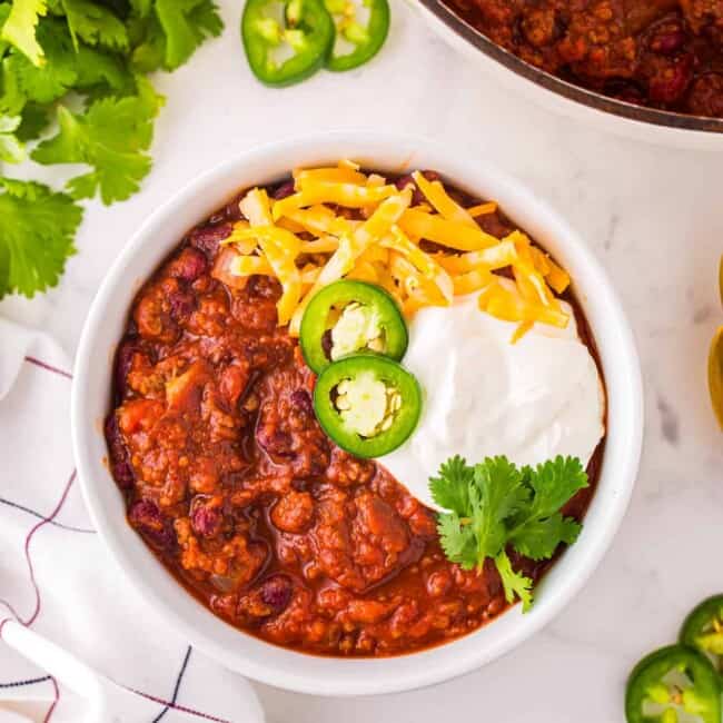 overhead view of chili con carne in a white bowl with cheese, sour cream, jalapenos, and cilantro.