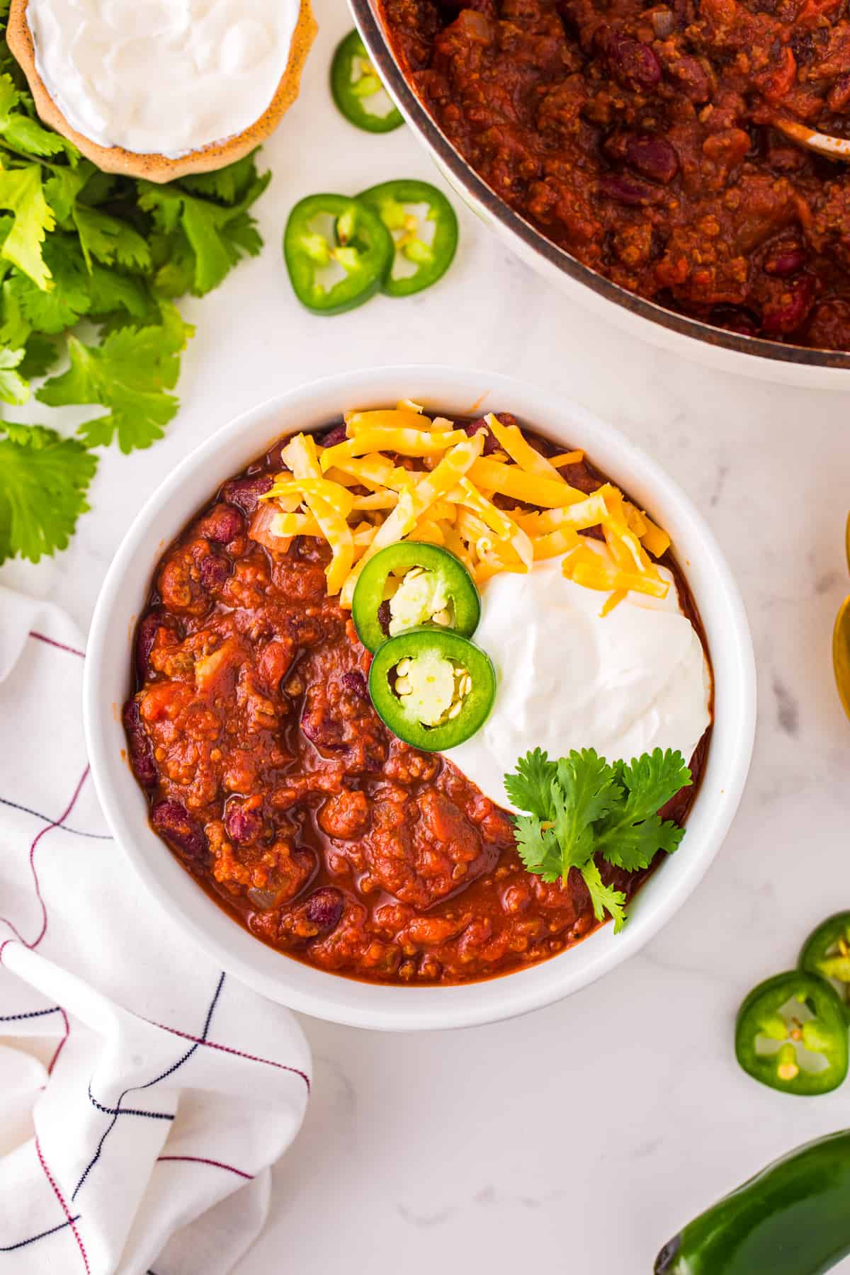 overhead view of chili con carne in a white bowl with cheese, sour cream, jalapenos, and cilantro.