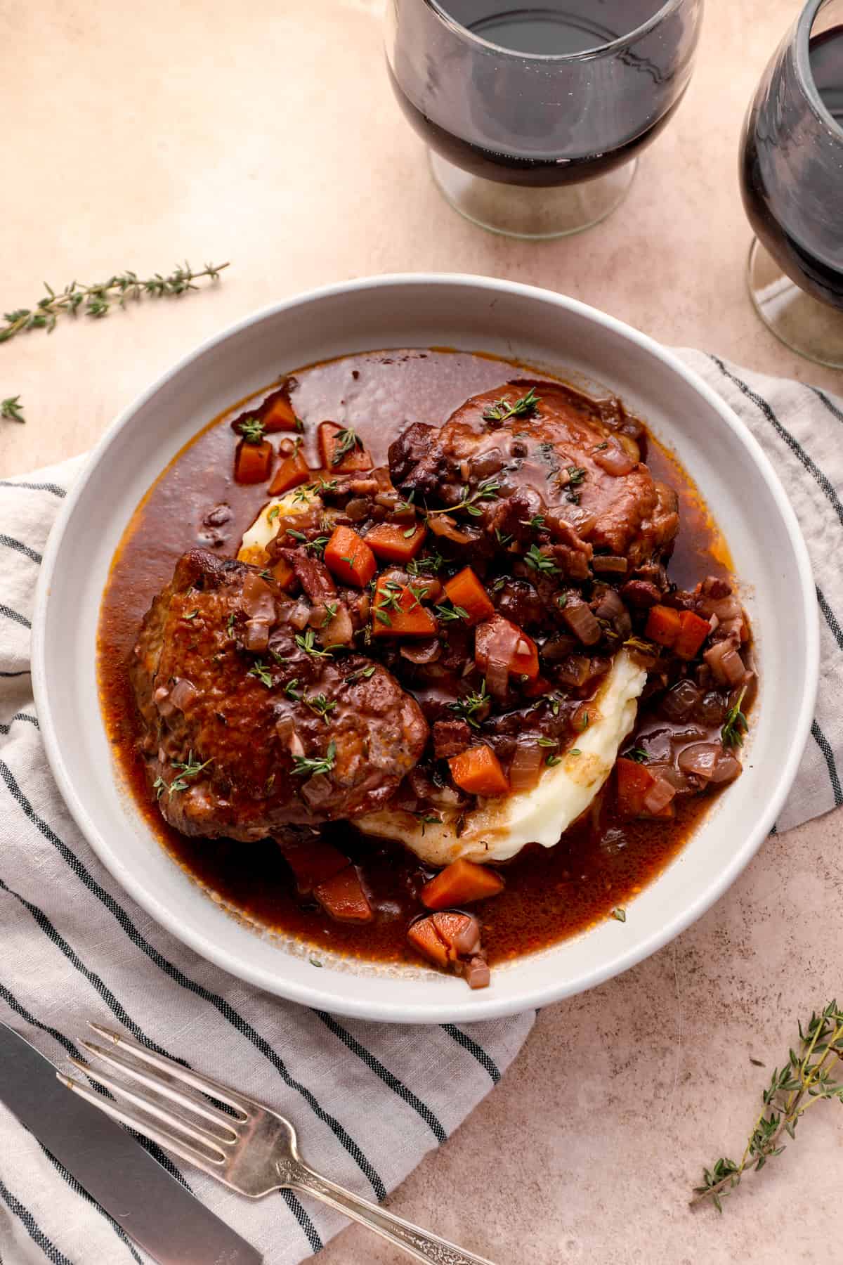 bowl of coq au vin on a table, with a dish towel, fork, and glasses of red wine