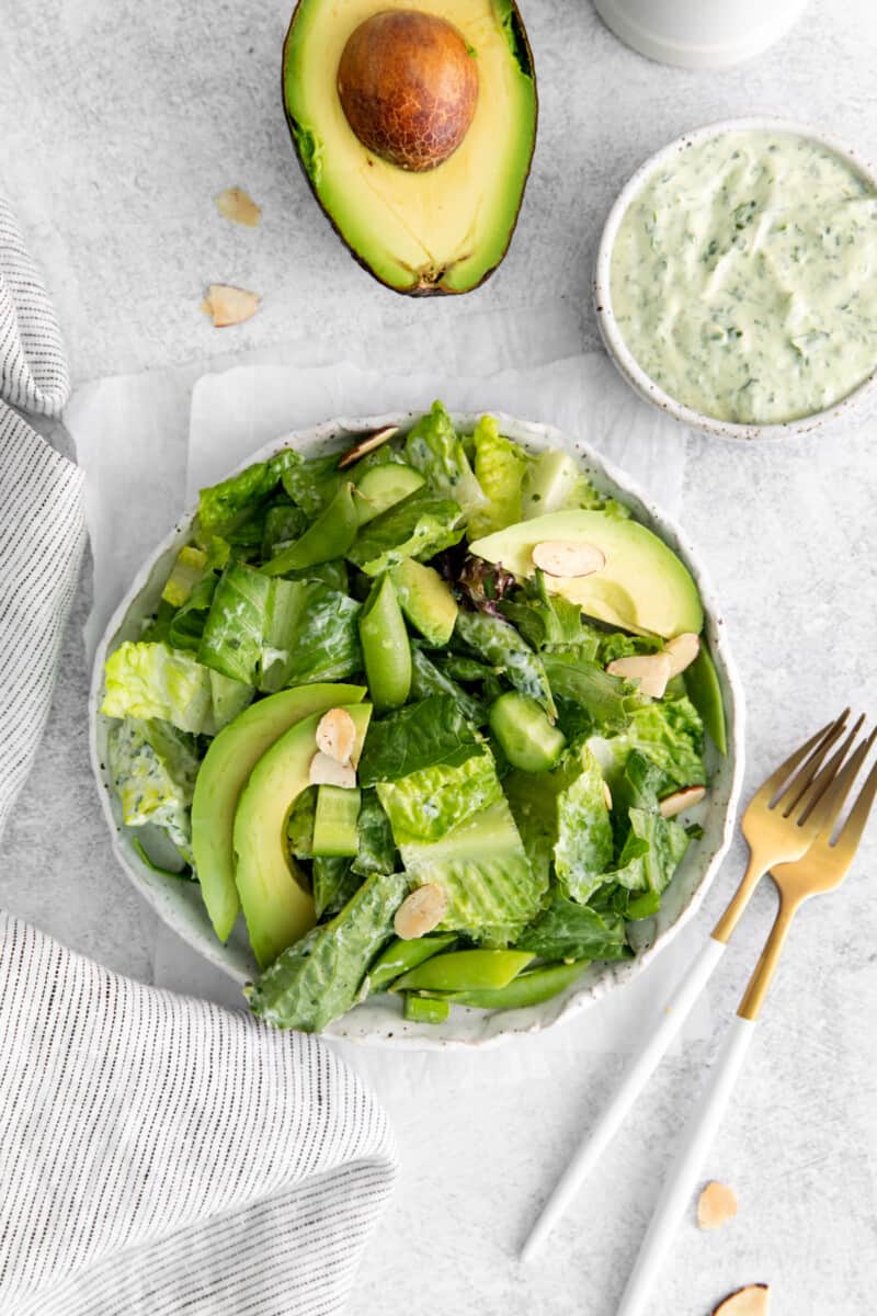 a green goddess salad on the table, arranged with forks, half an avocado, and a small bowl of dressing