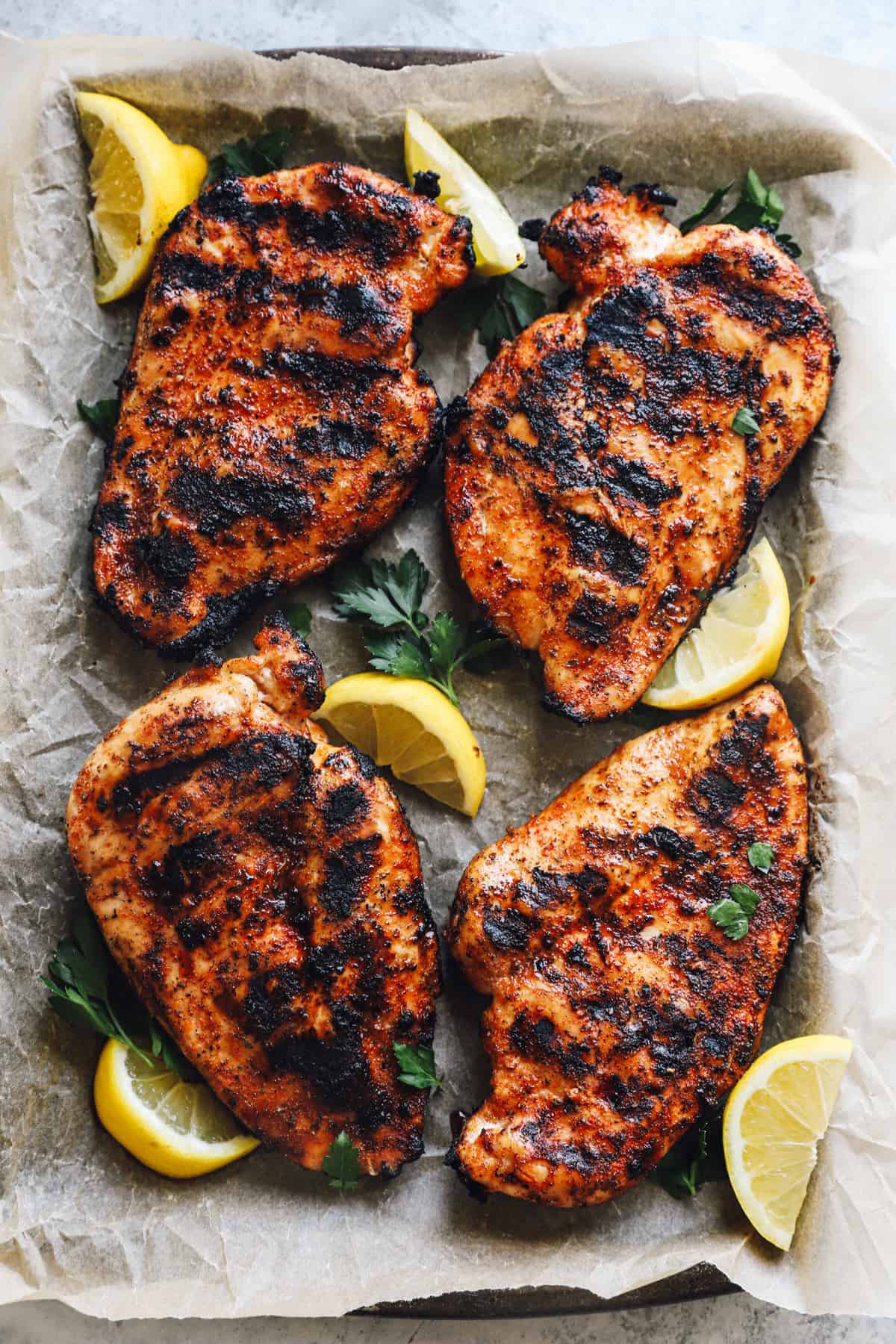 four pieces of grilled chicken breast, laying on a parchment-lined baking tray, surrounded by lemon slices
