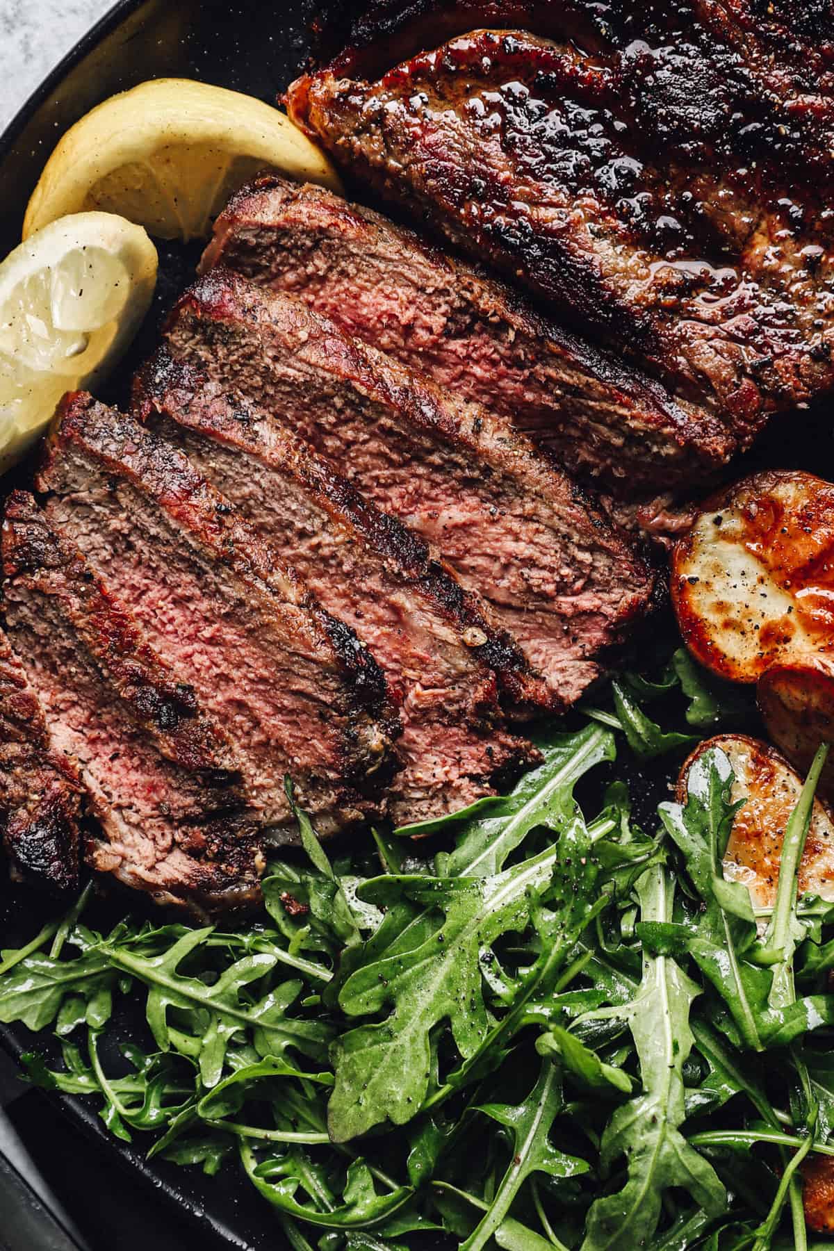 Sliced grilled ribeye with potatoes, arugula, and lemon wedges.