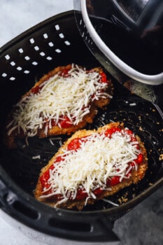 2 air fried chicken breasts in the basket of an air fryer topped with marinara sauce and cheese.