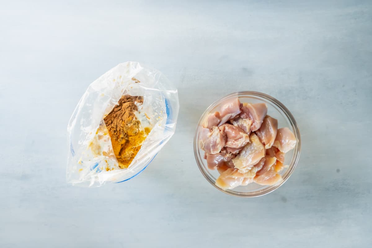 seasonings for indian butter chicken in a ziplock bag next to a glass bowl full of cut chicken.