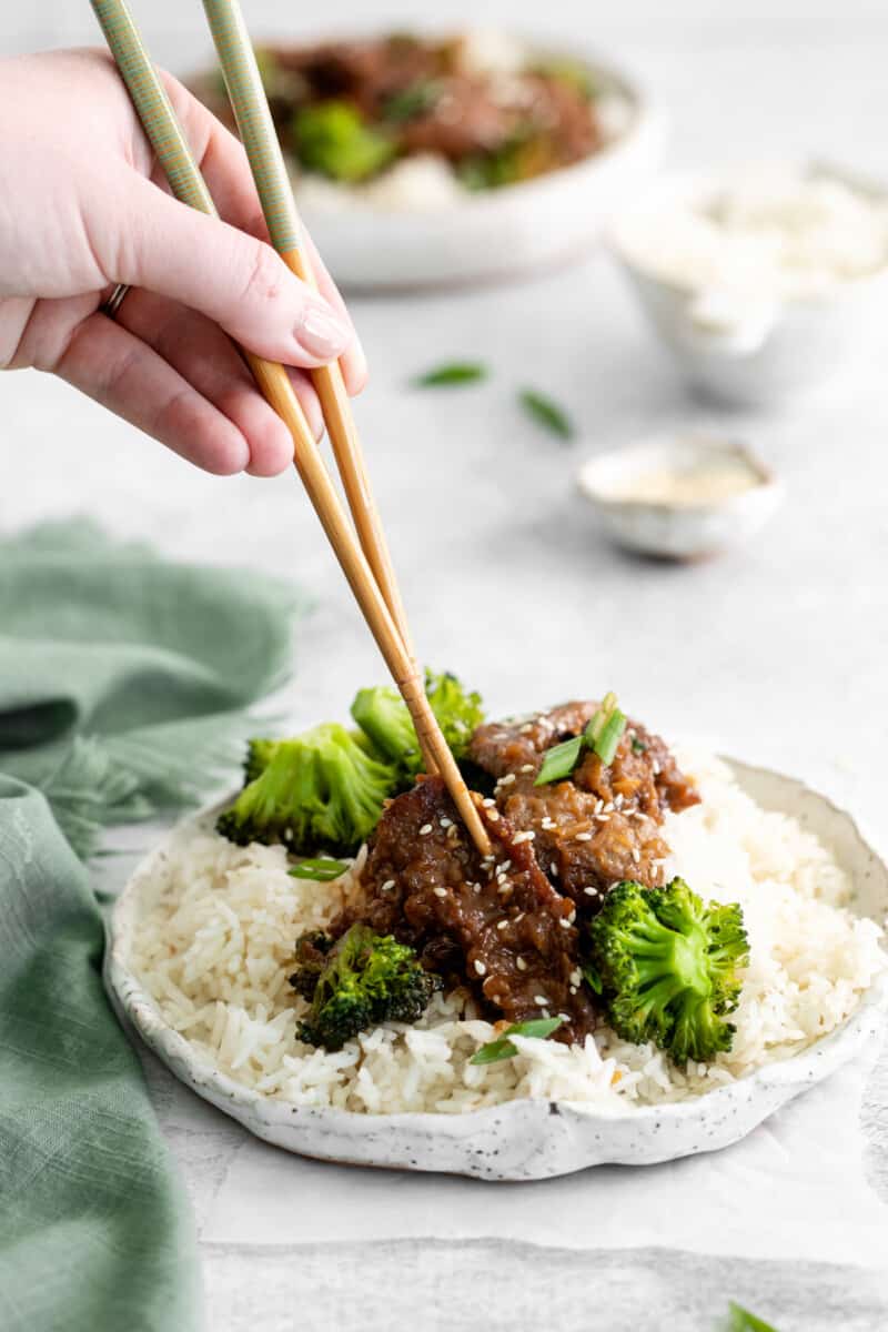 a hand using chopsticks to grab a piece of instant pot mongolian beef from a white plate.
