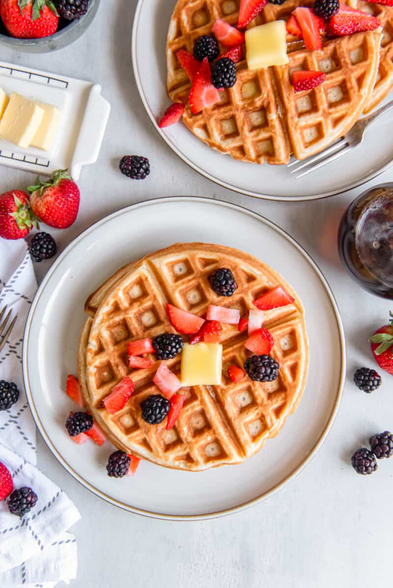 overhead view of belgian waffles on white plates with fruit and butter.