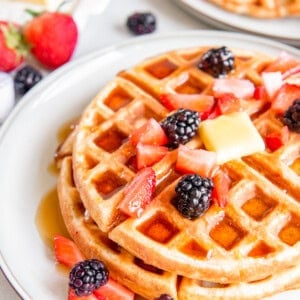 close up of 2 belgian waffles on a white plate with butter and fruit.