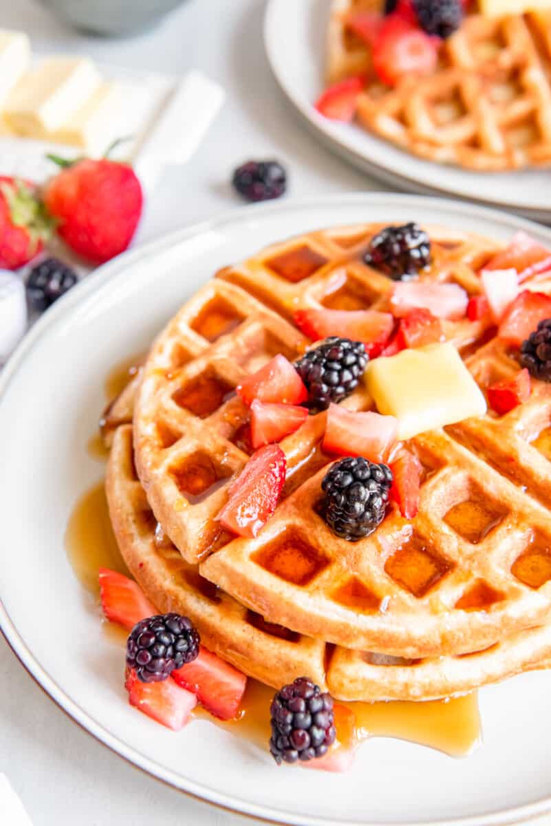 close up of 2 belgian waffles on a white plate with butter and fruit.