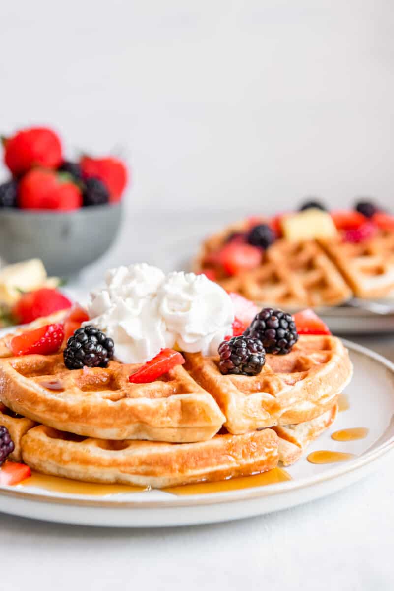 side view of 2 belgian waffles on a white plate with whipped cream and fruit.