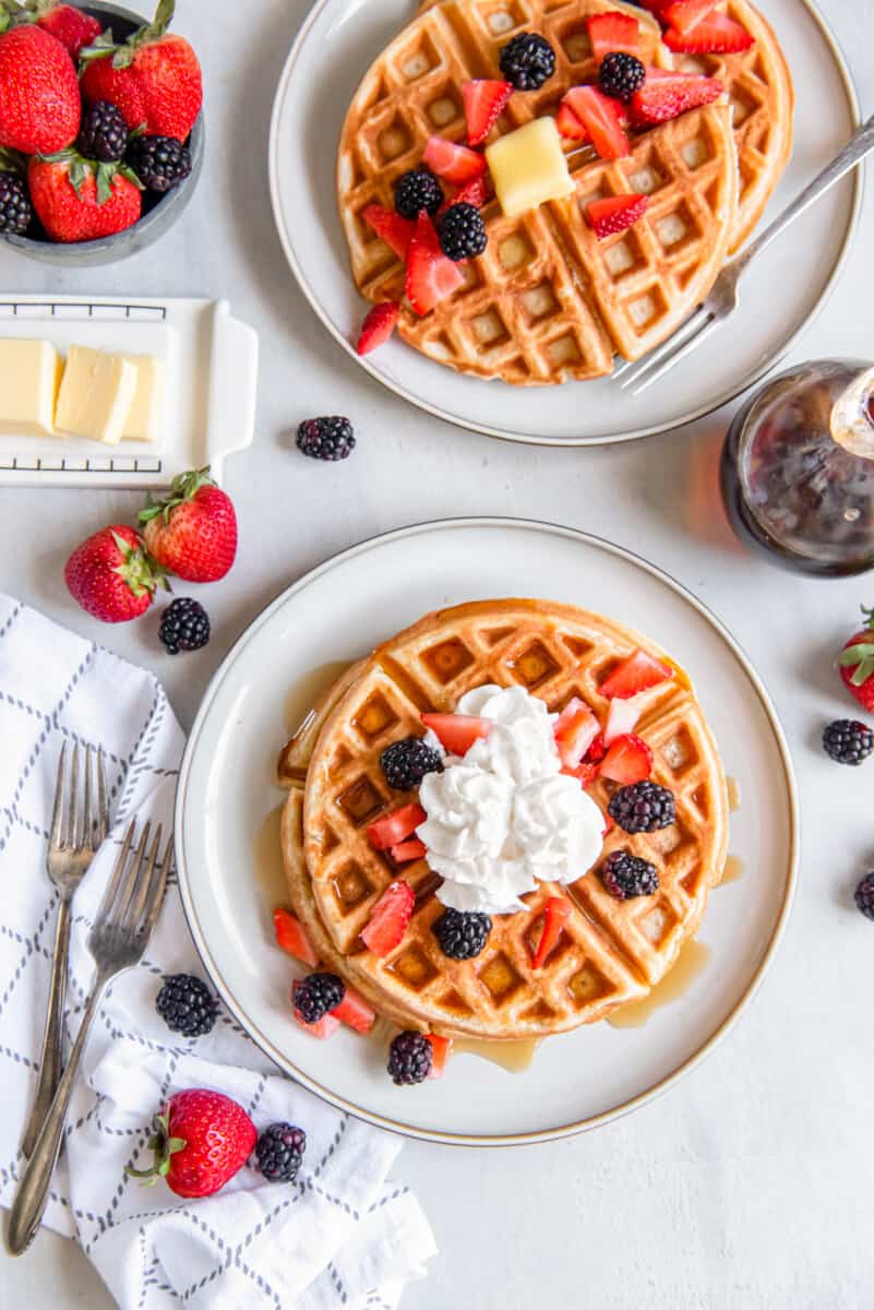 overhead view of belgian waffles on white plates with butter, whipped cream, and fruit,