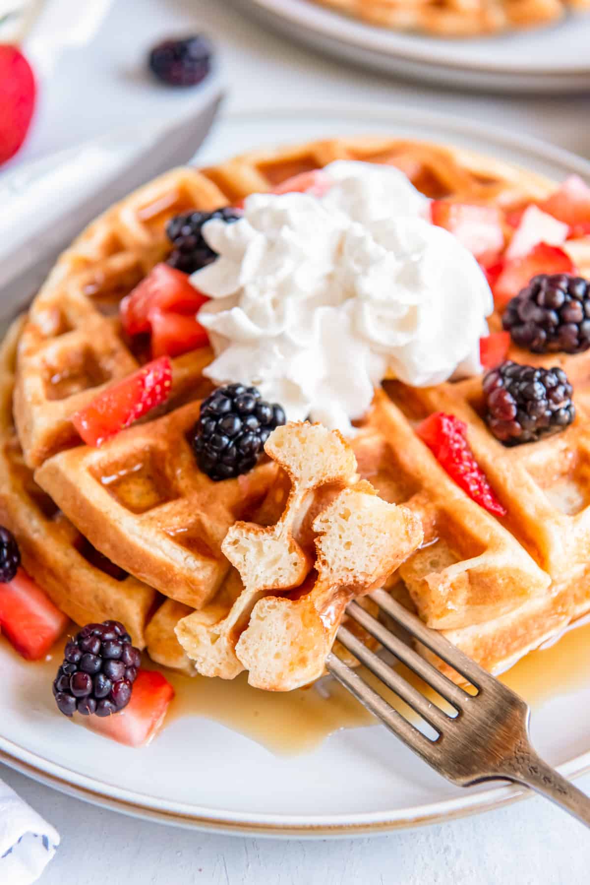 a bite of belgian waffle on a fork in front of a stack of 2 belgian waffles on a white plate with whipped cream and fruit.