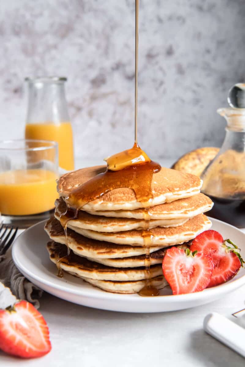 a tall stack of buttermilk pancakes on a white plate with butter, syrup, and a halved strawberry.