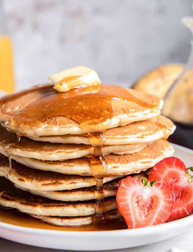 close up of a tall stack of buttermilk pancakes on a white plate with butter, syrup, and a halved strawberry.