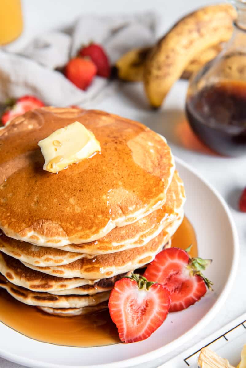 close up of a tall stack of buttermilk pancakes on a white plate with butter, syrup, and a halved strawberry.