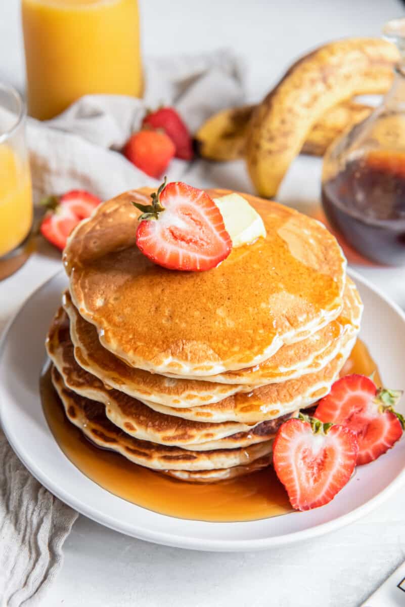 a tall stack of buttermilk pancakes on a white plate with butter, syrup, and a halved strawberry.