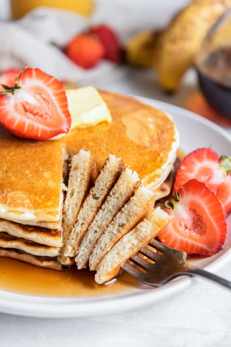 a forkful of buttermilk pancakes resting on a plate of buttermilk pancakes with syrup, butter, and a halved strawberry.