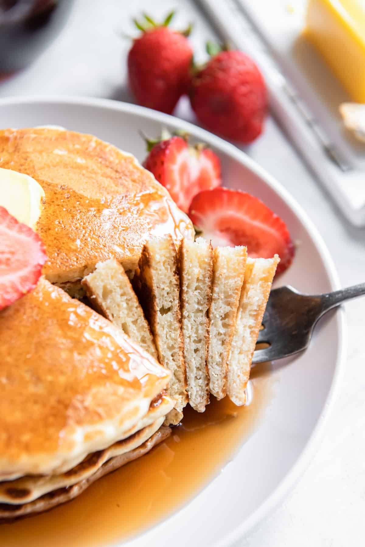 a forkful of buttermilk pancakes resting on a plate of buttermilk pancakes with syrup, butter, and a halved strawberry.