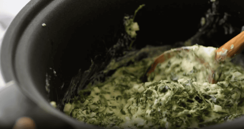 Using a crockpot, a person is stirring a bowl of spinach dip.