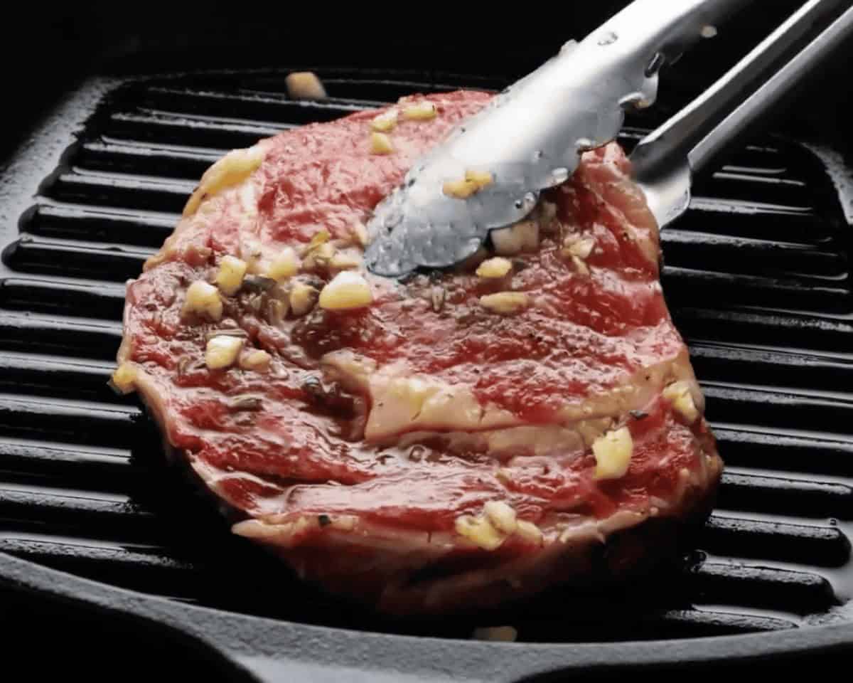 Ribeye steak covered in marinade on a grill being flipped with tongs.