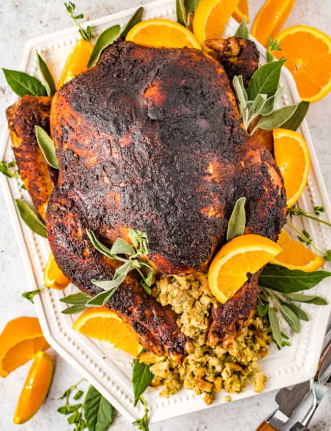 A rubbed whole turkey on a white serving tray with stuffing, sage, and orange slices.