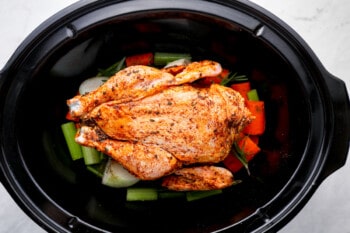 a whole chicken on a bed of chopped vegetables and herbs in a crockpot.