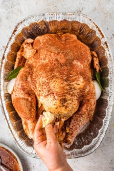overhead view of a hand buttering a raw herb brined turkey in an aluminum roasting pan.