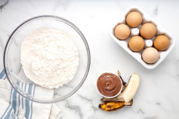 dry ingredients for banana nutella muffins in a glass bowl next to a half-dozen eggs, a peeled banana, and nutella.