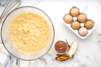banana nutella muffin batter in a glass bowl next to a half-dozen eggs, a peeled banana, and nutella.