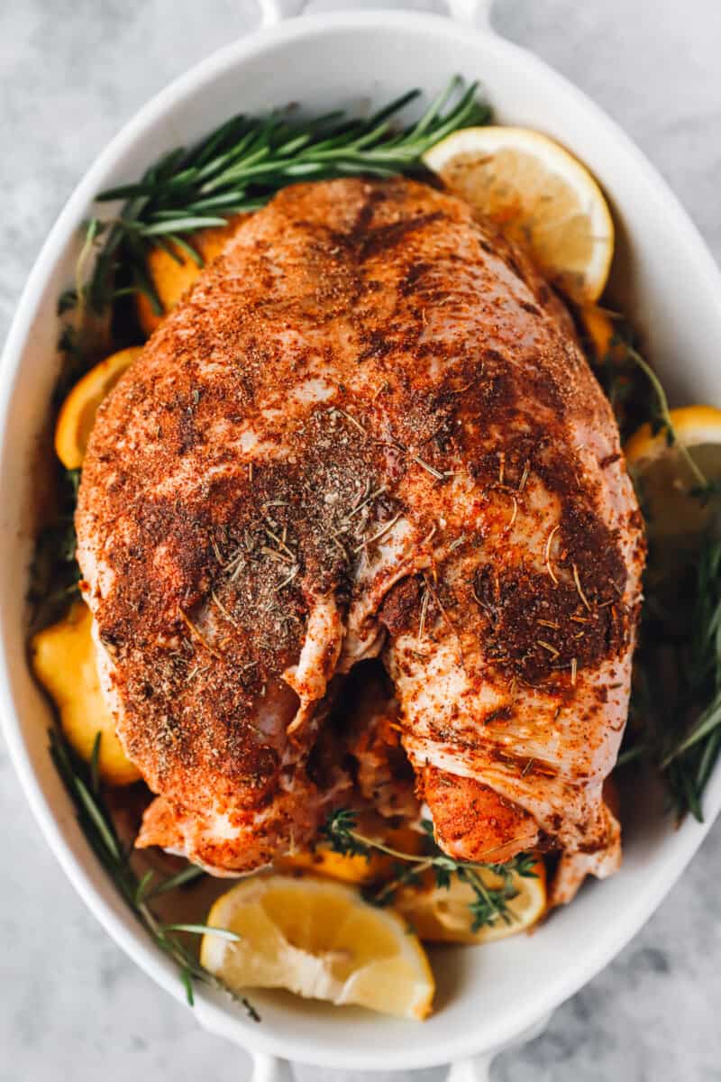 overhead view of a rubbed turkey breast in a white casserole dish with herbs and lemon wedges.