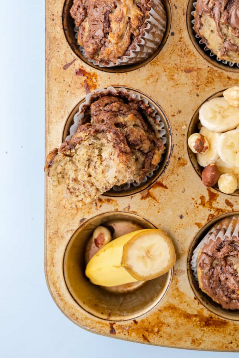 close up of a bitten banana nutella muffin resting on a muffin tin with a halved banana and banana slices.