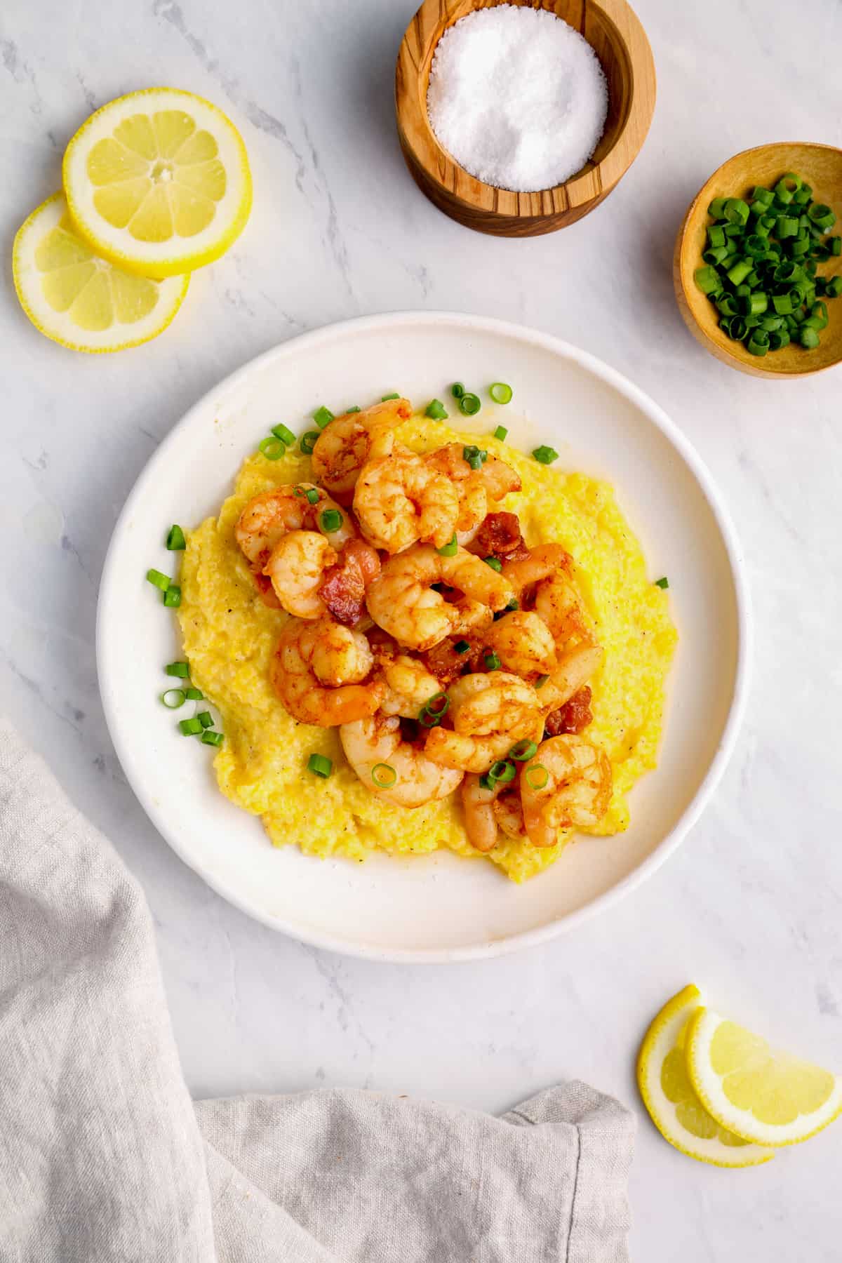 a plate of shrimp and grits next to small bowls of chives and salt, and lemon slices