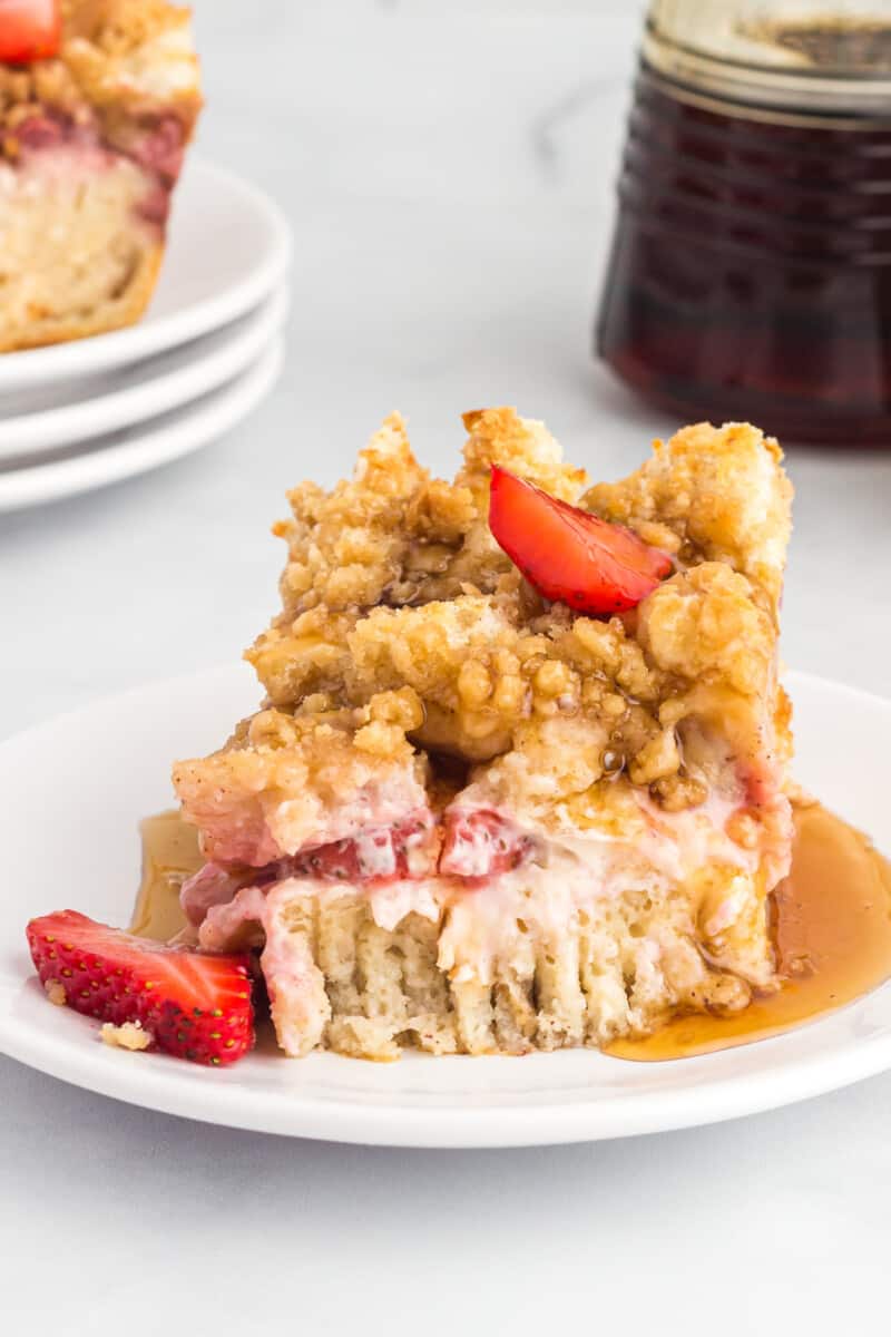 a slice of strawberry toast casserole on a plate, topped with syrup and fresh strawberries